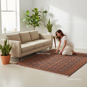 Puede incluir: Una alfombra rectangular marrón con un patrón geométrico está colocada sobre un suelo de color claro. La alfombra está en un salón, con un sofá beige, plantas en macetas y una mujer arrodillada cerca de la alfombra.