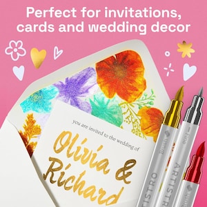 K&ouml;nnte beinhalten: Eine Hochzeitseinladung mit goldfarbenem Schriftzug in einem blumengef&uuml;tterten Umschlag. Drei metallische Kunststifte in Gold, Silber und Rot werden auf einem rosa Hintergrund angezeigt. Die Einladung lautet "Olivia & Richard".