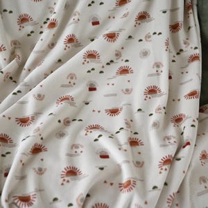 May include: Cream-colored fabric with a repeating pattern of stylized suns, clouds, and small green shapes. The suns are in shades of orange and brown. The fabric is draped, showing folds and texture.