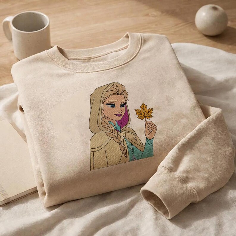 Puede incluir: Una sudadera de cuello redondo en color beige con un bordado de un personaje rubio con peinado trenzado y capa con capucha, sosteniendo una hoja de arce amarilla. El personaje tiene ojos azules, mechones morados y viste una prenda de color verde azulado.
