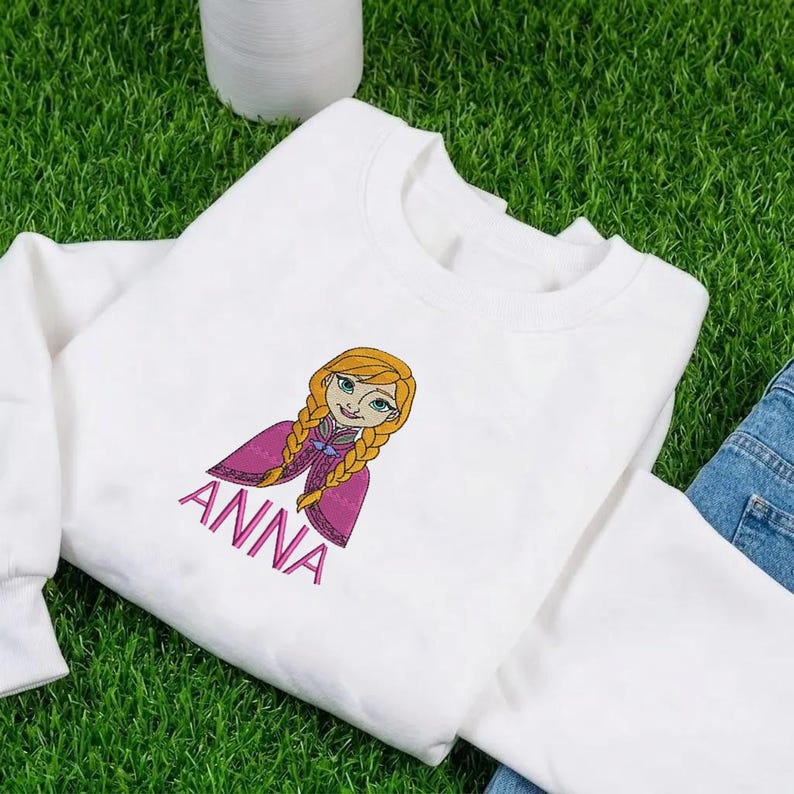 K&ouml;nnte beinhalten: Ein wei&szlig;es Langarm-Sweatshirt mit einer Stickerei einer Zeichentrickfigur mit geflochtenem blondem Haar, die einen magentafarbenen Umhang mit violetten Akzenten tr&auml;gt. Der Name ANNA ist darunter in rosa Gro&szlig;buchstaben eingestickt.