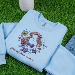 May include: A light blue sweatshirt featuring an embroidered design of a mermaid with brown hair, a purple seashell bra, and surrounded by starfish and flowers. The text "Part of Your World" is embroidered below the design.