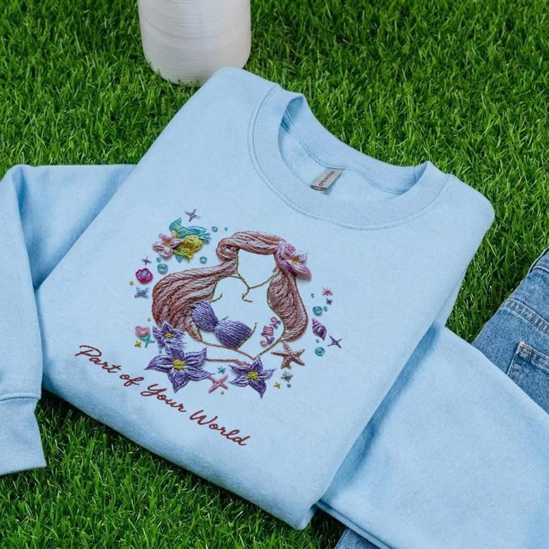 May include: A light blue sweatshirt featuring an embroidered design of a mermaid with brown hair, a purple seashell bra, and surrounded by starfish and flowers. The text "Part of Your World" is embroidered below the design.