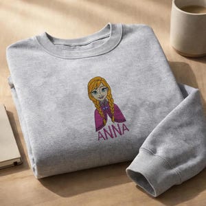 K&ouml;nnte beinhalten: Ein hellgraues Sweatshirt mit Rundhalsausschnitt und einer gestickten Illustration einer Zeichentrickfigur mit geflochtenem blonden Haar und violettem Umhang, &uuml;ber dem in pinkfarbenem Garn gestickten Namen ANNA auf der Brust.