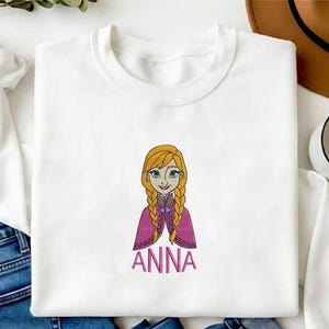 K&ouml;nnte beinhalten: Ein wei&szlig;es Langarm-Sweatshirt mit einer Stickerei einer Zeichentrickfigur mit geflochtenem blondem Haar, die einen magentafarbenen Umhang &uuml;ber einem dunklen Oberteil tr&auml;gt. Der Name ANNA ist darunter in rosa Gro&szlig;buchstaben eingestickt.