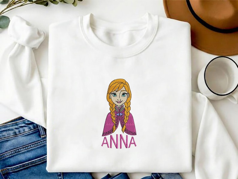 K&ouml;nnte beinhalten: Ein wei&szlig;es Langarm-Sweatshirt mit einer Stickerei einer Zeichentrickfigur mit geflochtenem blondem Haar, die einen magentafarbenen Umhang &uuml;ber einem dunklen Oberteil tr&auml;gt. Der Name ANNA ist darunter in rosa Gro&szlig;buchstaben eingestickt.