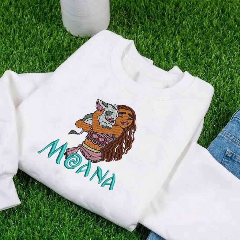 May include: A white sweatshirt featuring an embroidered graphic of a long-haired character holding a small pig. Below the character, the name MOANA is embroidered in teal thread. The sweatshirt has long sleeves and a crew neckline.