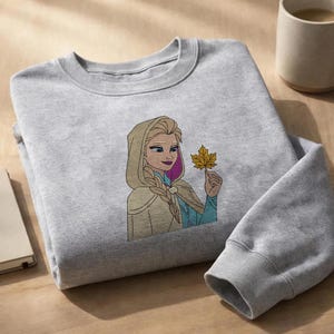 Puede incluir: Una sudadera gris claro de cuello redondo con un bordado de un personaje rubio con peinado trenzado, que viste una capa beige con capucha y una prenda azul, sosteniendo una hoja de arce amarilla de oto&ntilde;o en una mano.