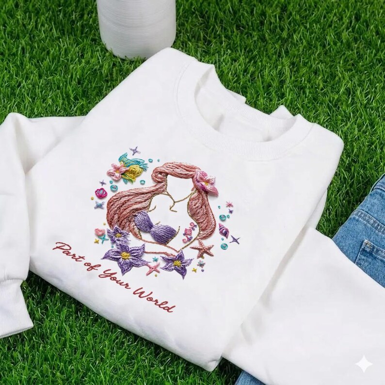 May include: White sweatshirt featuring an embroidered design of a mermaid with auburn hair, a purple seashell bra, and surrounded by flowers and starfish. The text "Part of Your World" is embroidered in red. The sweatshirt is made of a soft material.