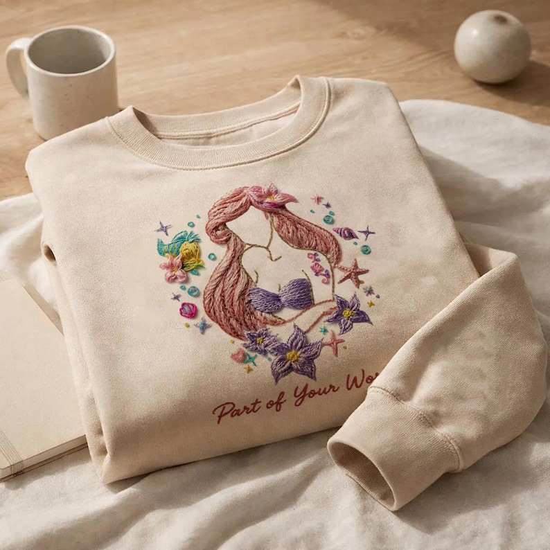 May include: Beige sweatshirt with an embroidered design of a mermaid with pink hair and a purple seashell bra. The design includes flowers, starfish, and the text "Part of Your Wo". The sweatshirt is folded on a white surface.