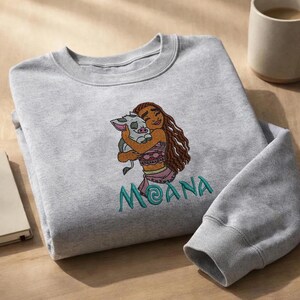 May include: A light gray crewneck sweatshirt featuring an embroidered design of a long-haired character hugging a small pig. Below the embroidered illustration, the name MOANA is stitched in teal thread with a stylized letter O.