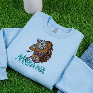 May include: A light blue crewneck sweatshirt featuring an embroidered graphic of a brown-haired character holding a small pig. The name MOANA is embroidered in teal thread below the character. The sweatshirt has long sleeves and a ribbed collar.
