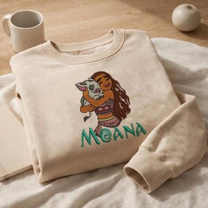 May include: A beige crewneck sweatshirt featuring an embroidered design of a long-haired character hugging a small piglet. Below the characters, the name MOANA is embroidered in teal green thread with a stylized letter A.
