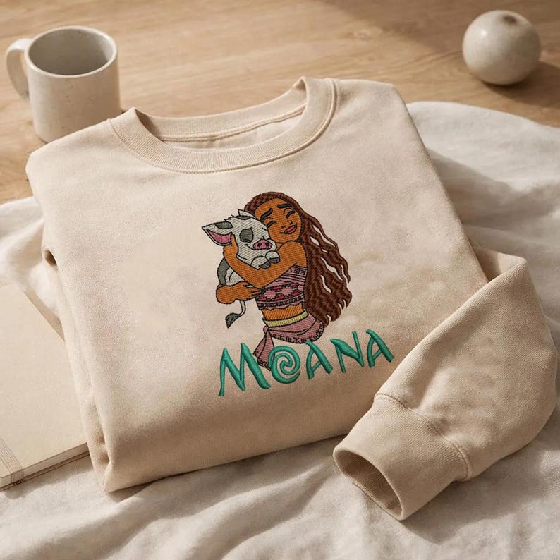 May include: A beige crewneck sweatshirt featuring an embroidered design of a long-haired character hugging a small piglet. Below the characters, the name MOANA is embroidered in teal green thread with a stylized letter A.