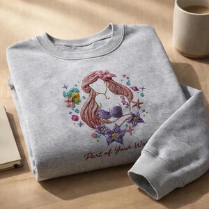 May include: A gray sweatshirt featuring an embroidered design of a mermaid with red hair and a purple seashell bra, surrounded by flowers and starfish. The text "Part of Your World" is below the design.
