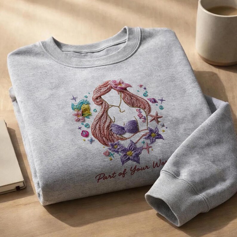 May include: A gray sweatshirt featuring an embroidered design of a mermaid with red hair and a purple seashell bra, surrounded by flowers and starfish. The text "Part of Your World" is below the design.