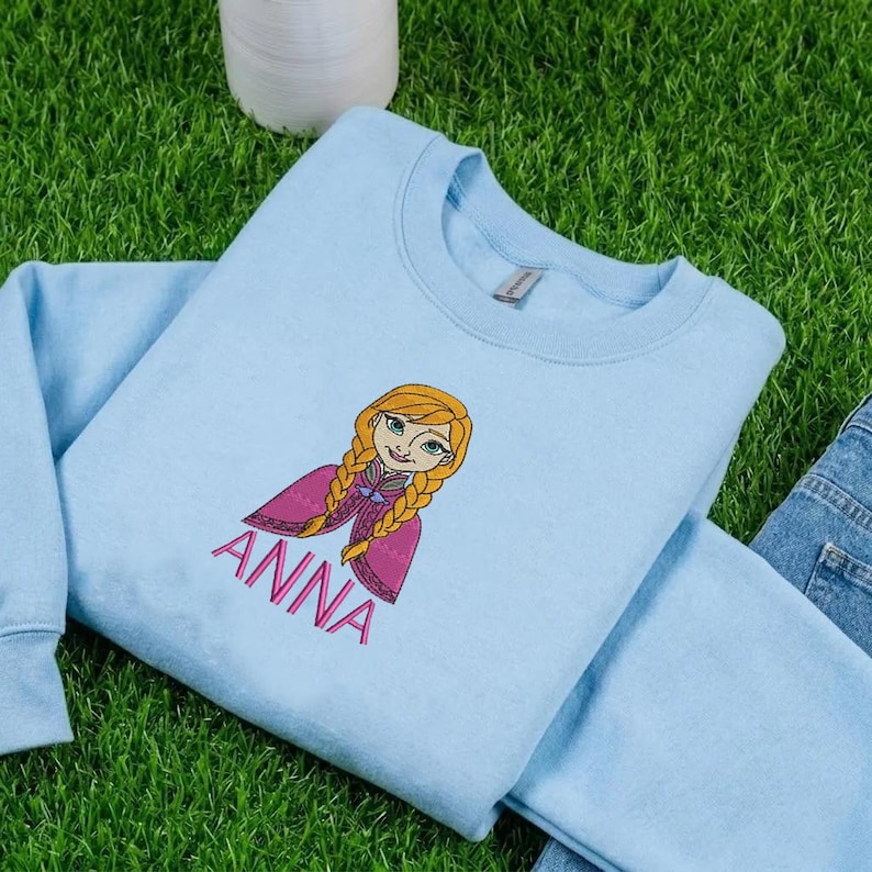 K&ouml;nnte beinhalten: Ein hellblaues Sweatshirt mit Rundhalsausschnitt und einer Stickerei der Figur Anna mit blonden Z&ouml;pfen und einem rosa Umhang. Der Name ANNA ist in rosa Gro&szlig;buchstaben direkt unter der Illustration der Figur eingestickt.
