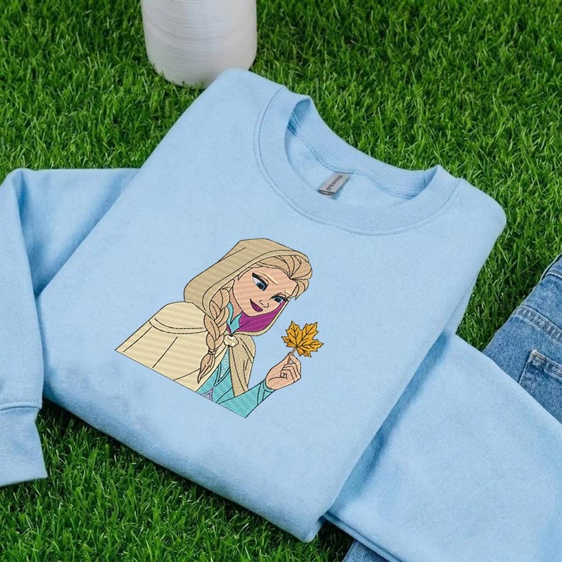 Puede incluir: Una sudadera de cuello redondo en color azul claro con un bordado que muestra a un personaje rubio con peinado trenzado y capa con capucha de color beige, sosteniendo una hoja de oto&ntilde;o amarilla. El dise&ntilde;o presenta costuras detalladas.