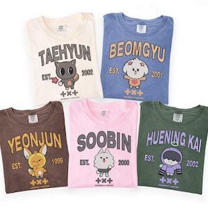 Puede incluir: Cinco camisetas dobladas en crema, azul, marrón, rosa y verde. Cada camiseta presenta un nombre, un personaje de dibujos animados y el texto "EST." con un año. Las camisetas tienen un aspecto vintage descolorido.