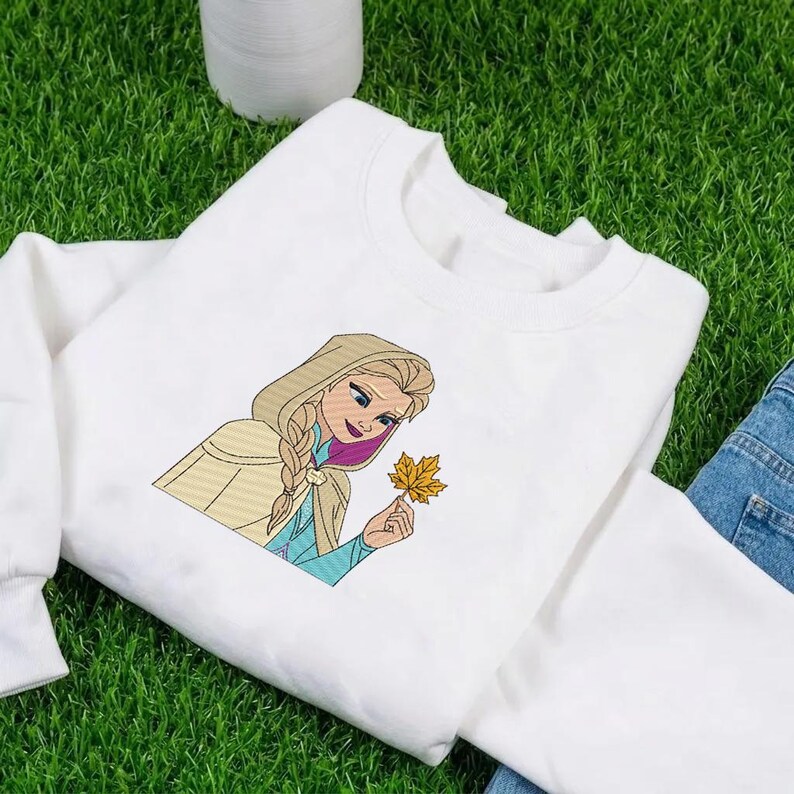 Puede incluir: Una sudadera blanca de manga larga con un gr&aacute;fico bordado de un personaje rubio con peinado trenzado, que lleva una capa beige con capucha y sostiene una hoja de arce amarilla. El personaje tiene mechones de pelo morados y una prenda azul.