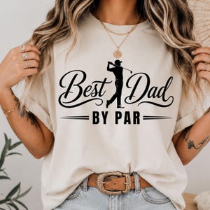 May include: A cream-colored t-shirt featuring a black graphic print of a golfer in mid-swing. The text reads "Best Dad" in a cursive script above the golfer and "BY PAR" in a bold, sans-serif font below, accented by horizontal lines.