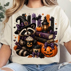 May include: A cream-colored t-shirt featuring a graphic of a raccoon wearing a purple-patterned witch hat and scarf. The raccoon holds a jack-o'-lantern mug with whipped cream. Nearby are stacked books, a lantern, and a large carved pumpkin.