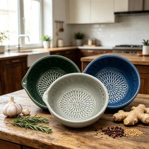 Op de afbeelding: Drie keramische keukenschalen in groen, grijs en blauw, elk met een bloemmotief in het midden. De kommen staan op een houten oppervlak met knoflook, rozemarijn, gember en peperkorrels. De kommen hebben een handvat aan de zijkant.