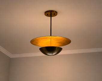 May include: A modern, two-tiered pendant light fixture. The top tier is a large, circular, gold-colored disc, and the bottom tier is a dark, bowl-shaped element. The fixture hangs from a bronze-colored ceiling mount and rod.