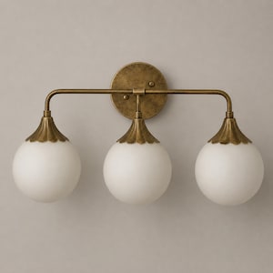 May include: A three-light wall sconce featuring a circular antique brass backplate and a horizontal bar supporting three downward-facing white frosted glass globe shades, each topped with a scalloped brass cap and a curved metal arm.