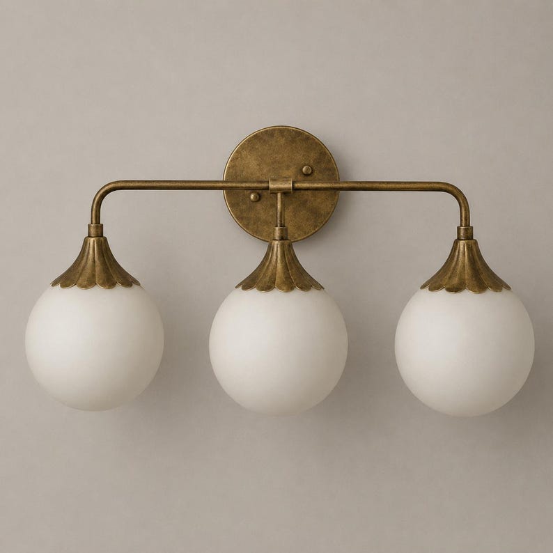 May include: A three-light wall sconce featuring a circular antique brass backplate and a horizontal bar supporting three downward-facing white frosted glass globe shades, each topped with a scalloped brass cap and a curved metal arm.