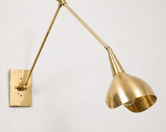 May include: A brass wall-mounted lamp with an adjustable arm and a conical shade. The lamp is mounted on a rectangular base and extends outward with a jointed arm. The shade is open at the bottom, revealing the light bulb.
