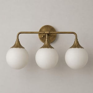 May include: A three-light wall sconce featuring a distressed antique brass finish. The fixture has a horizontal bar supporting three spherical, frosted white glass globes, each attached to a scalloped metal cap that connects to the frame.