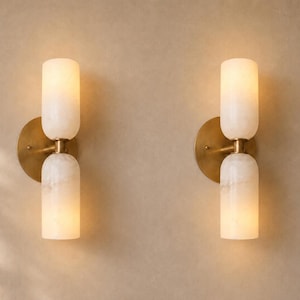 May include: A pair of wall sconces featuring two cylindrical, translucent white stone shades stacked vertically. Each light is mounted on a circular, brushed brass backplate, with a central brass connector holding the stone shades in place.