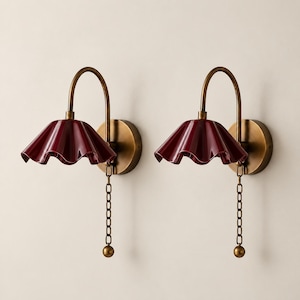 Könnte beinhalten: Ein Paar Wandleuchten mit einem tiefroten, gewellten Keramikschirm, der an einem geschwungenen, messingfarbenen Metallarm befestigt ist. Jede Lampe verfügt über eine runde Wandhalterung und eine passende Zugkette aus Messing mit einer kleinen Kugel.