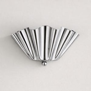 Op de afbeelding: Een gepolijste, zilverkleurige wandlamp met een geschulpte, waaiervormige vormgeving. De lamp heeft een glad, reflecterend oppervlak en een klein, bolvormig detail aan de onderkant. Het ontwerp doet denken aan een schelp.