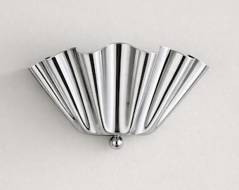 Könnte beinhalten: Eine polierte, silberfarbene Wandleuchte mit einem wellenförmigen, fächerartigen Design. Die Leuchte hat eine glatte, reflektierende Oberfläche und ein kleines, kugelförmiges Detail an der Unterseite. Das Design erinnert an eine Muschel.