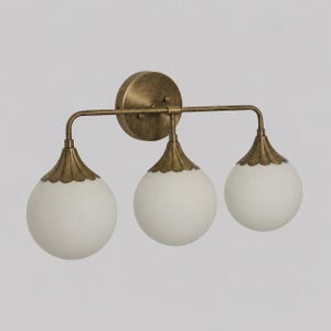 May include: A three-light wall sconce featuring a circular antique brass backplate and a horizontal bar supporting three spherical, frosted white glass globes. Each globe is capped with a scalloped, antique brass metal fixture.