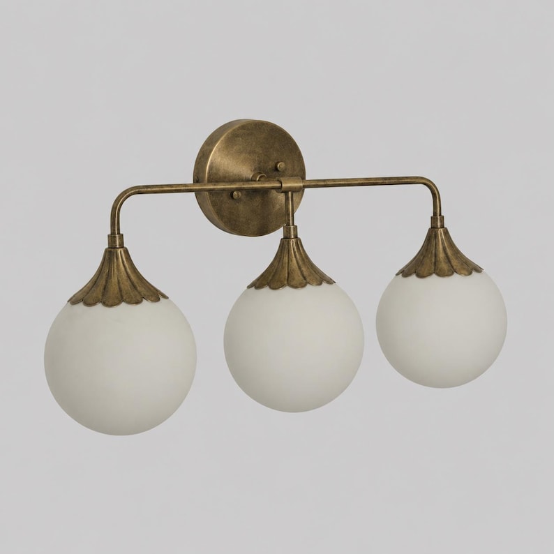 May include: A three-light wall sconce featuring a circular antique brass backplate and a horizontal bar supporting three spherical, frosted white glass globes. Each globe is capped with a scalloped, antique brass metal fixture.