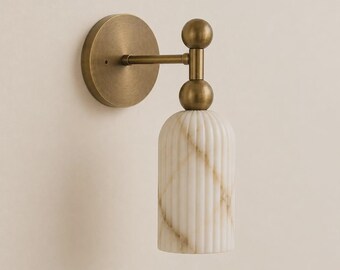 May include: A wall sconce featuring a circular antique brass backplate and a horizontal arm connected to a vertical post with two spherical brass accents. The light fixture includes a cylindrical, vertically ribbed white marble shade with tan veining.