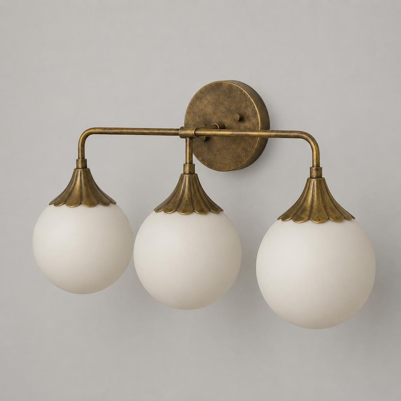May include: A three-light wall sconce featuring a circular antique brass backplate and a horizontal bar. Three spherical white frosted glass globes are suspended from the bar, each topped with a scalloped brass cap that connects to the fixture.