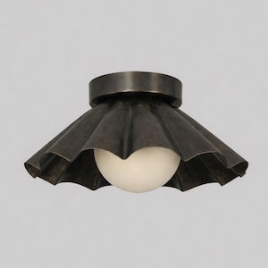 May include: A bronze-colored ceiling light fixture with a ruffled shade and a round, white light bulb. The shade has a scalloped edge and a circular base. The fixture is designed to be mounted flush against the ceiling.