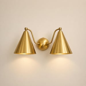 May include: A brass wall sconce featuring two conical shades. The shades extend outwards from a central sphere, connected by curved arms. The sconce is mounted on a light-coloured wall, creating a warm, inviting ambiance.