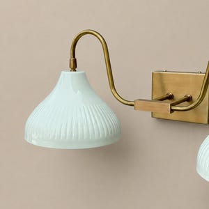 K&ouml;nnte beinhalten: Eine Wandleuchte mit zwei blass mintgr&uuml;nen, gerippten Keramikschirmen. Die Schirme sind an einem Arm aus geb&uuml;rstetem Messing und einer rechteckigen Wandplatte befestigt, was ein strukturiertes Design mit einer glatten, gl&auml;nzenden Oberfl&auml;che ergibt.
