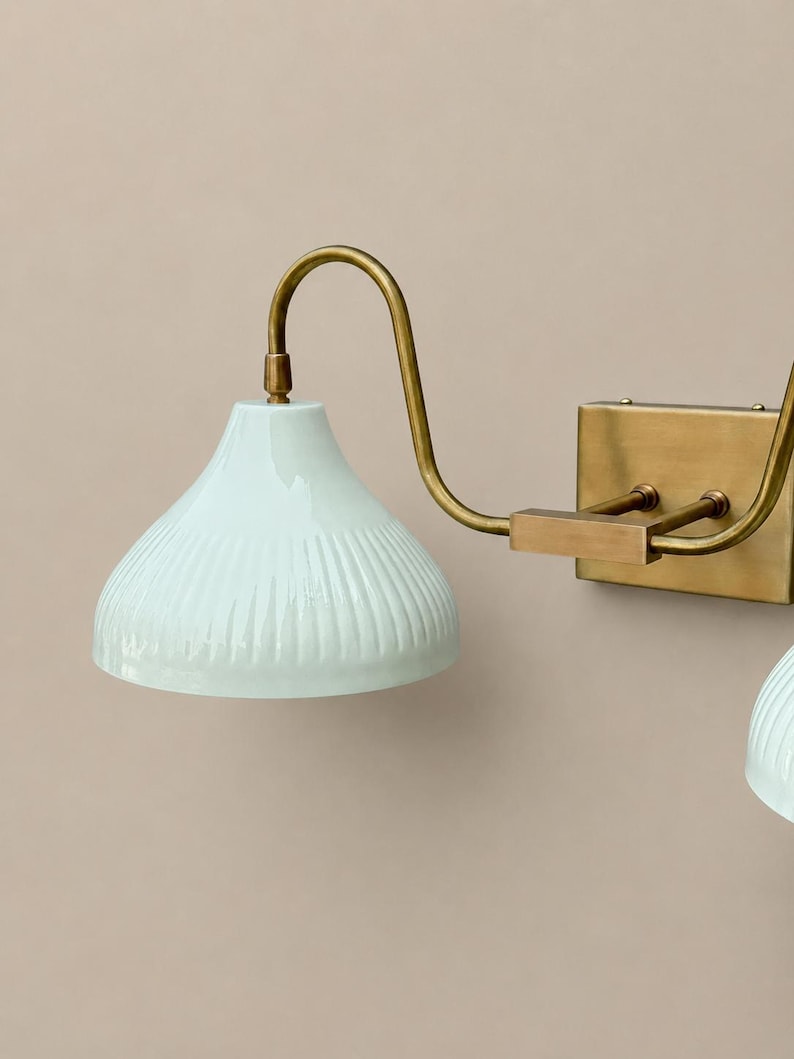 K&ouml;nnte beinhalten: Eine Wandleuchte mit zwei blass mintgr&uuml;nen, gerippten Keramikschirmen. Die Schirme sind an einem Arm aus geb&uuml;rstetem Messing und einer rechteckigen Wandplatte befestigt, was ein strukturiertes Design mit einer glatten, gl&auml;nzenden Oberfl&auml;che ergibt.