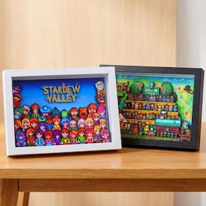 Könnte beinhalten: Zwei Schattenbox-Rahmen mit Stardew Valley Pixel-Art. Der linke Rahmen hat einen weißen Rand und zeigt eine Sammlung bunter Charakter-Sprites unter dem Text Stardew Valley. Der rechte Rahmen hat einen schwarzen Rand und zeigt eine detaillierte Szene eines Stadtfestes mit mehreren Charakteren, die auf gestuften Plattformen angeordnet sind.