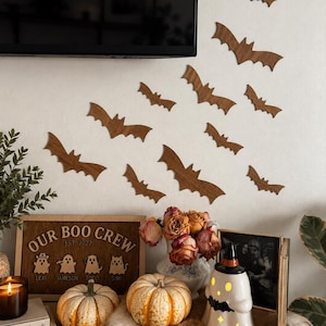 May include: A collection of ten brown wooden bat cutouts arranged on a wall. Below, a wooden sign reads "OUR BOO CREW EST 2022" with four ghost illustrations labeled Lexi, Jameson, Todd, and Simi, alongside two pumpkins and a ghost-shaped lamp.