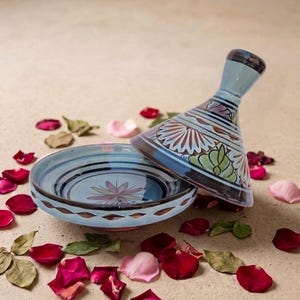 May include: A ceramic tagine with a conical lid and a shallow base. The piece features a light blue glaze decorated with hand-painted floral motifs in shades of brown, green, and white, accented by dark blue circular bands and a scalloped rim pattern.