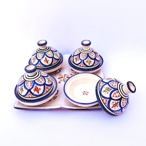Peut inclure: Ensemble de quatre petits plats à tajine en céramique avec couvercles, plus un plat ouvert, disposés sur un plateau rectangulaire. Les plats présentent une base blanche avec des motifs floraux bleus, rouges et verts. Les couvercles sont coniques avec une petite poignée.