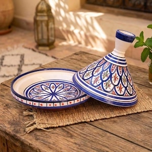 Peut inclure: Un tajine en céramique composé d'un couvercle conique et d'une base large, orné de motifs géométriques peints à la main en bleu, rouge et vert sur fond blanc. Le couvercle possède une poignée haute et étroite, et la base présente un motif central.