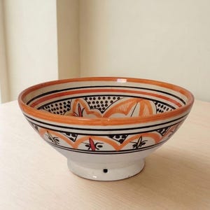 Peut inclure: Un bol en céramique à base blanche orné de motifs géométriques peints à la main en orange et noir. Le design comprend des lignes horizontales, des sections pointillées et des motifs floraux stylisés sur le bord intérieur et extérieur.